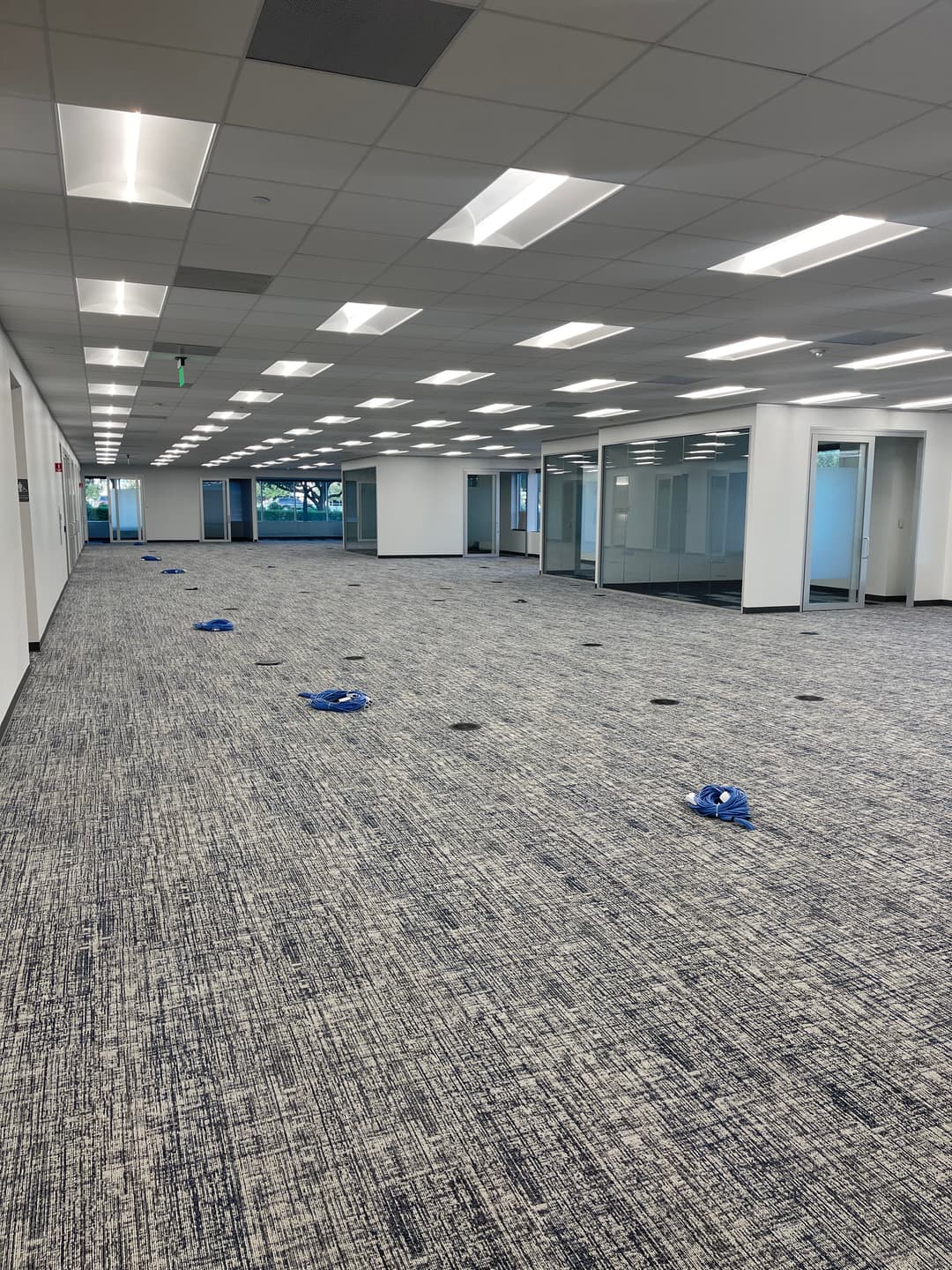 Empty office space with blue equipment on carpeted floor; large windows providing natural light.